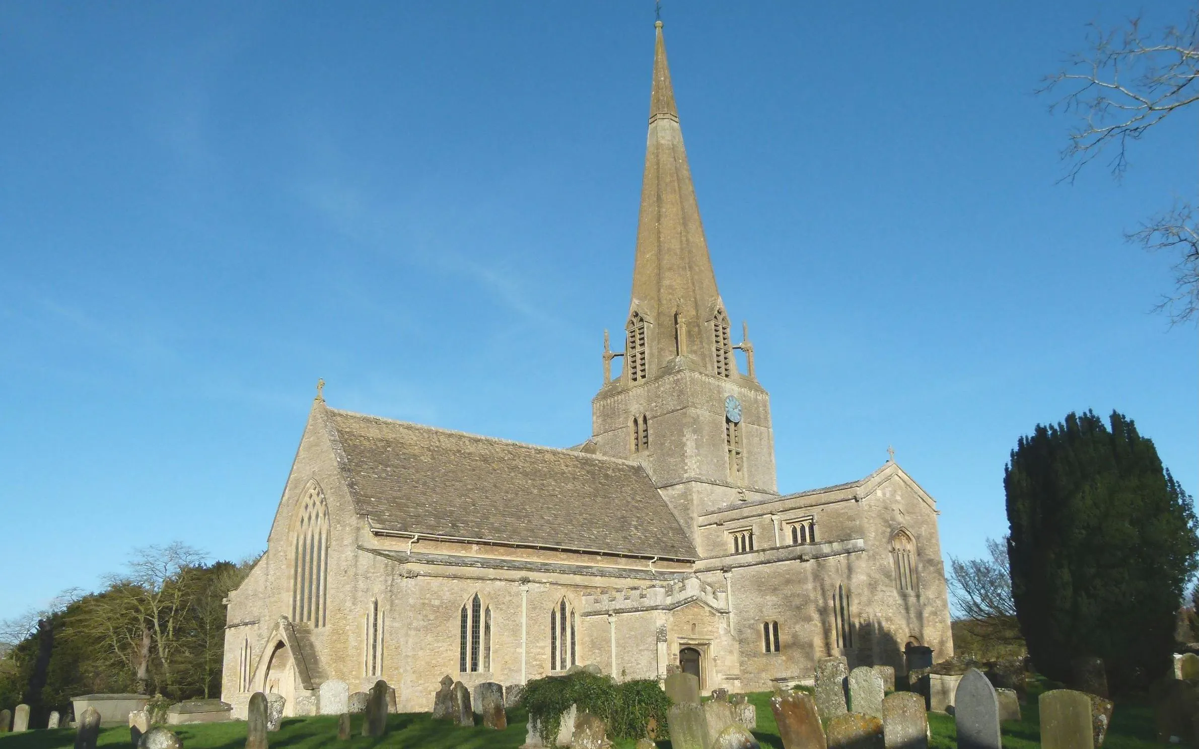 Bampton Church with tall spire and surrounding gravestones under clear blue sky.