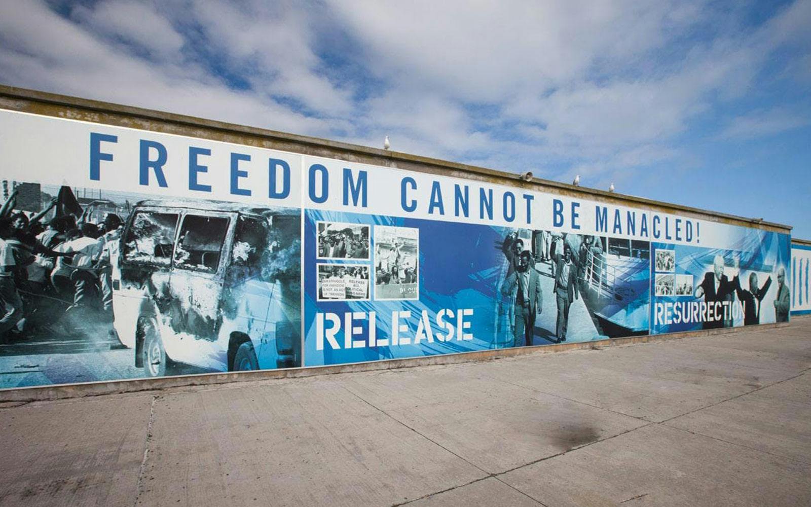 Mural with "Freedom Cannot Be Manacled" at Robben Island harbour, depicting historical events.