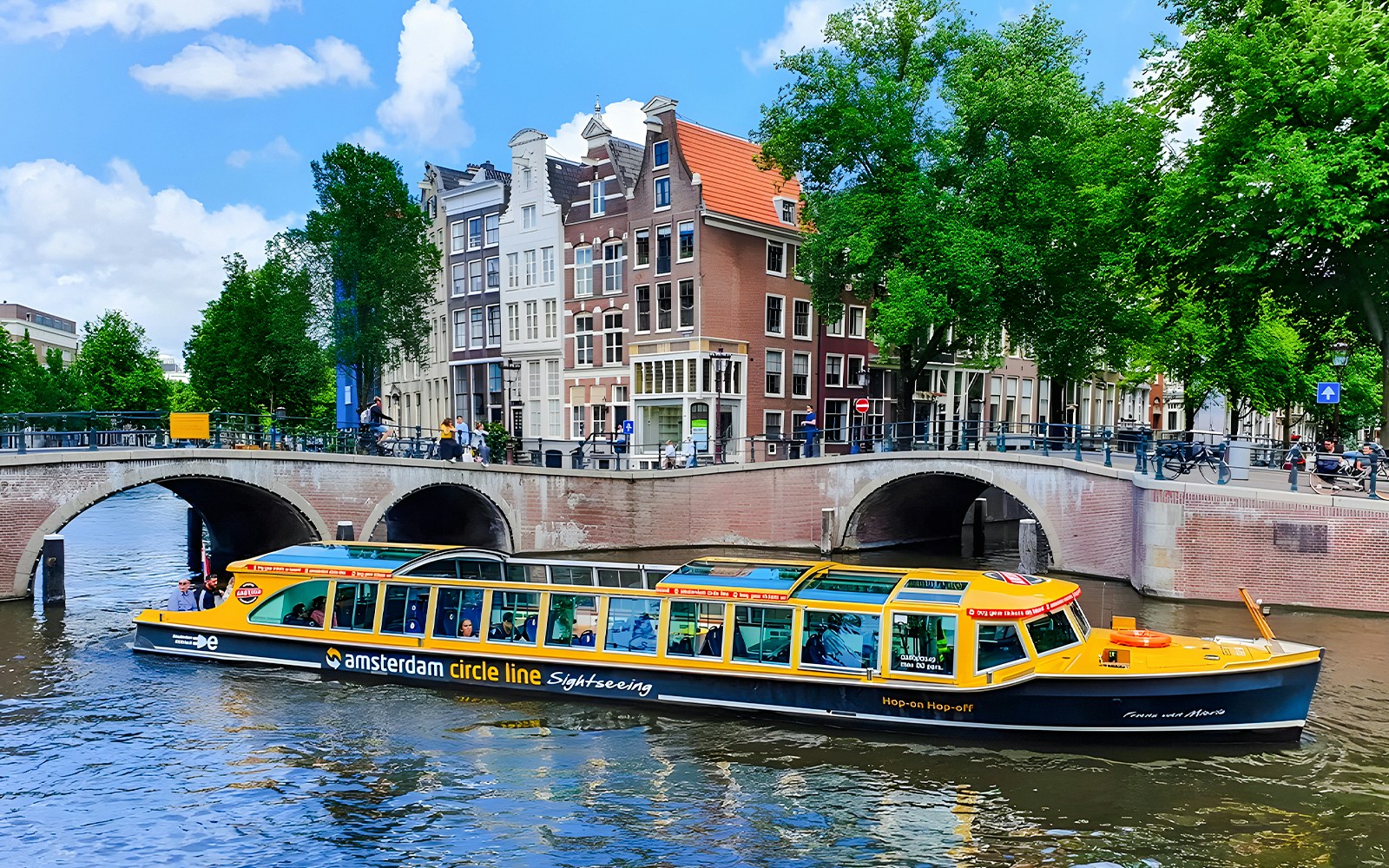 Amsterdam canal cruise boat passing under bridge with historic buildings in background.