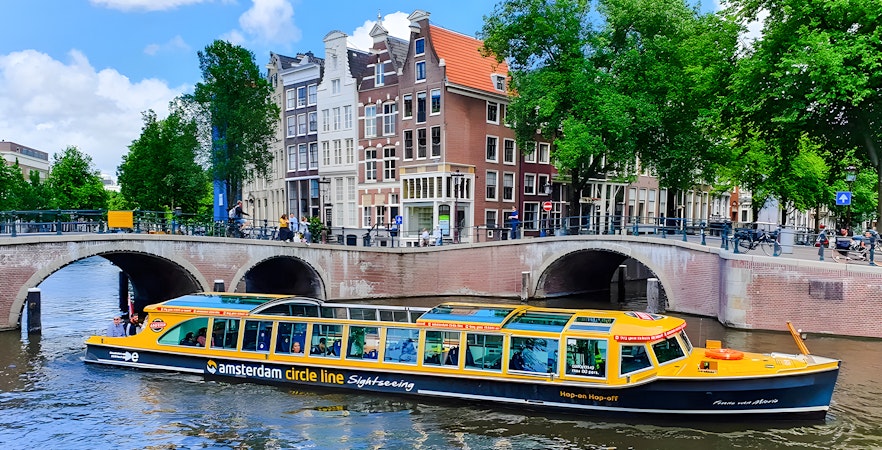 Amsterdam canal cruise boat passing under bridge with historic buildings in background.