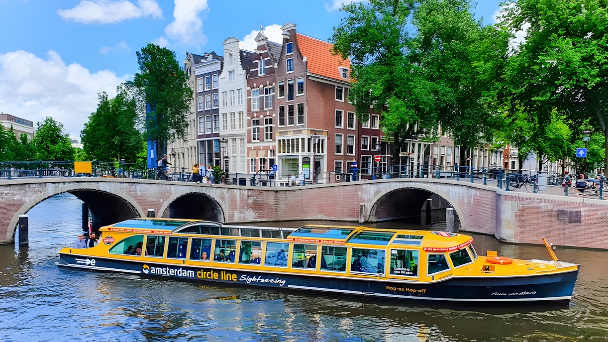Amsterdam canal cruise boat passing under bridge with historic buildings in background.