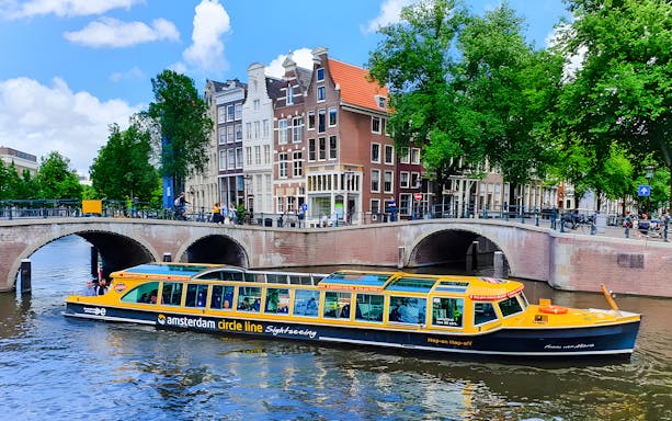 Amsterdam canal cruise boat passing under bridge with historic buildings in background.
