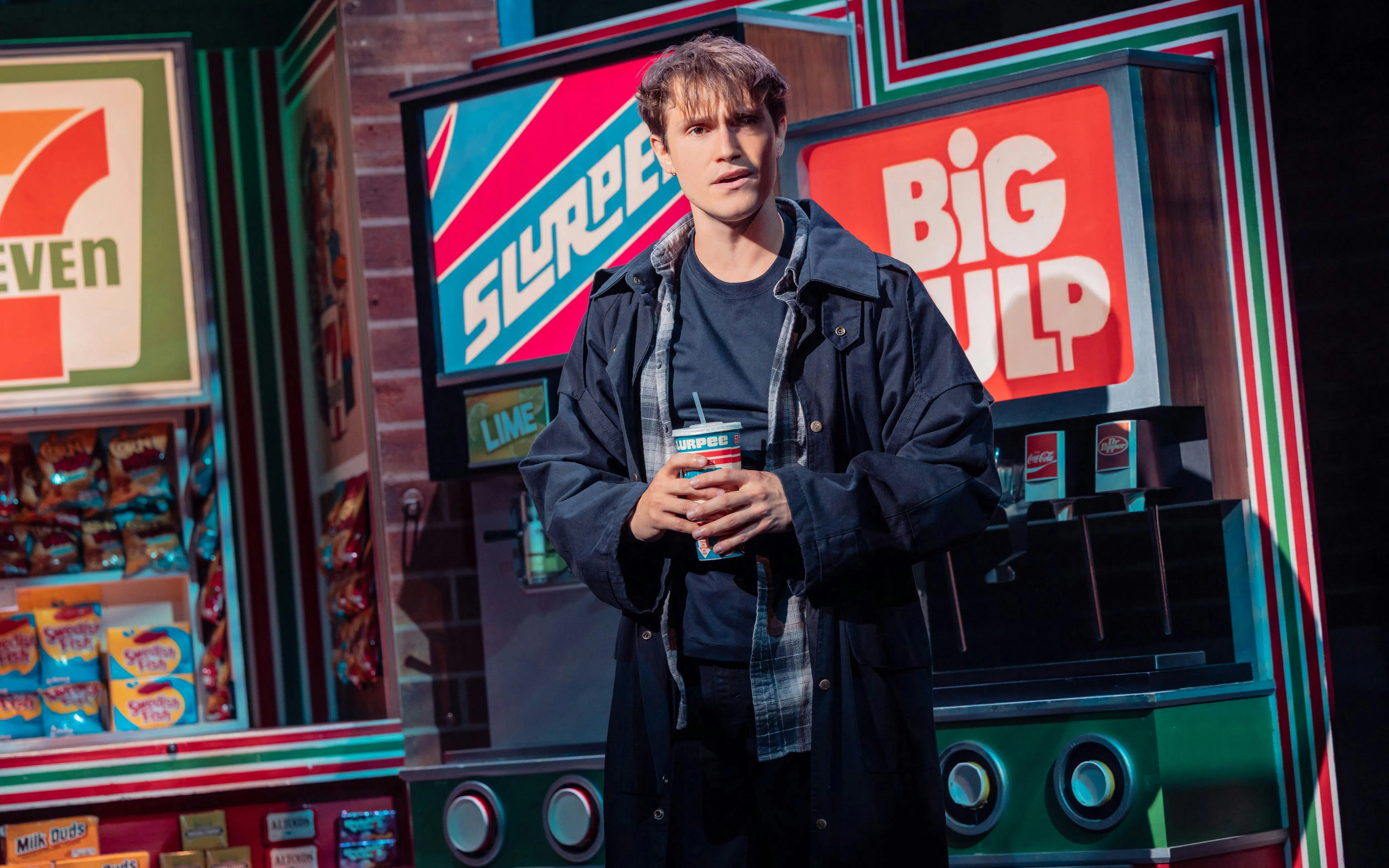 Actor on stage in Heathers The Musical holding a drink in a convenience store setting.