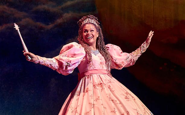 Performer in a pink gown holding a wand, portraying Mother Goose on stage.
