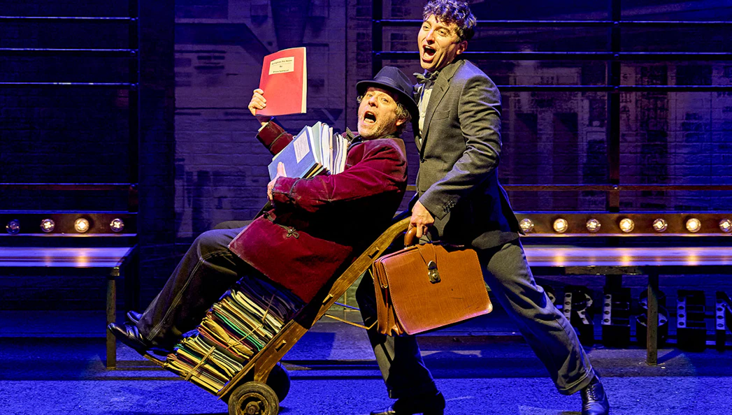 Two actors perform a comedic scene from "The Producers" on stage, one pushing a cart of scripts.