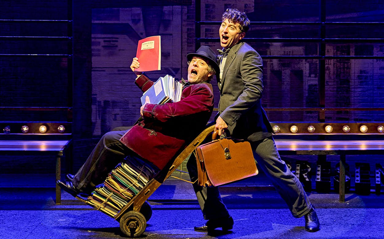Two actors perform a comedic scene from "The Producers" on stage, one pushing a cart of scripts.