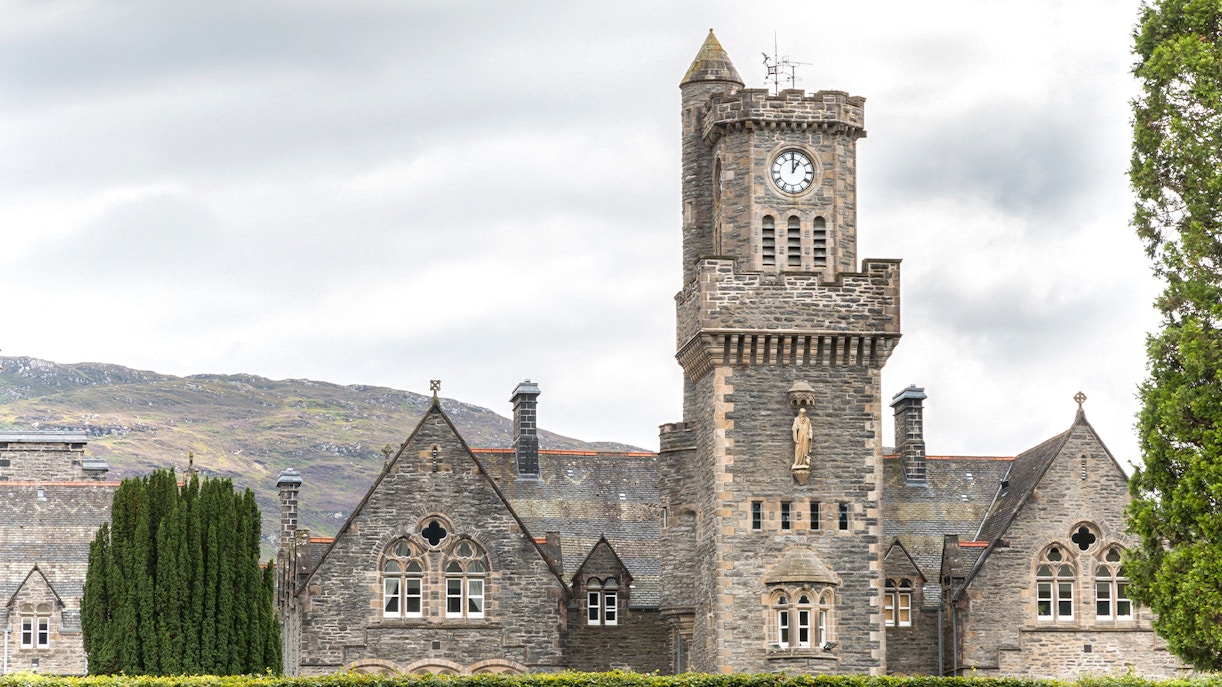 Clock tower and stone building in Fort Augustus, Scotland, with hills in the background.