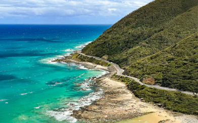 Aerial view of the Great Ocean Road winding along the coastline with turquoise waters and lush hills.