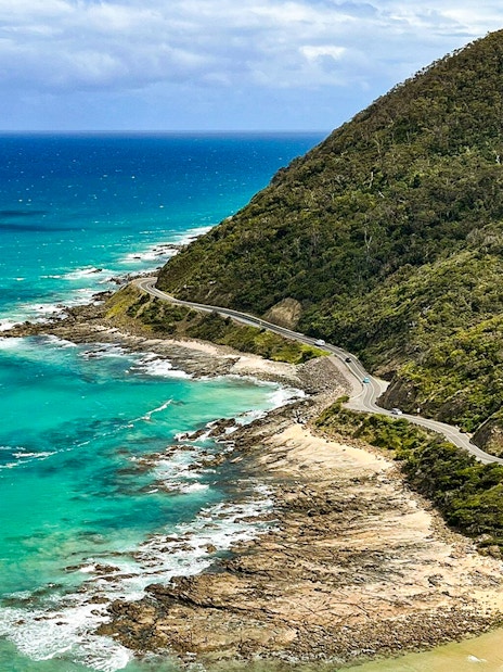 Aerial view of the Great Ocean Road winding along the coastline with turquoise waters and lush hills.