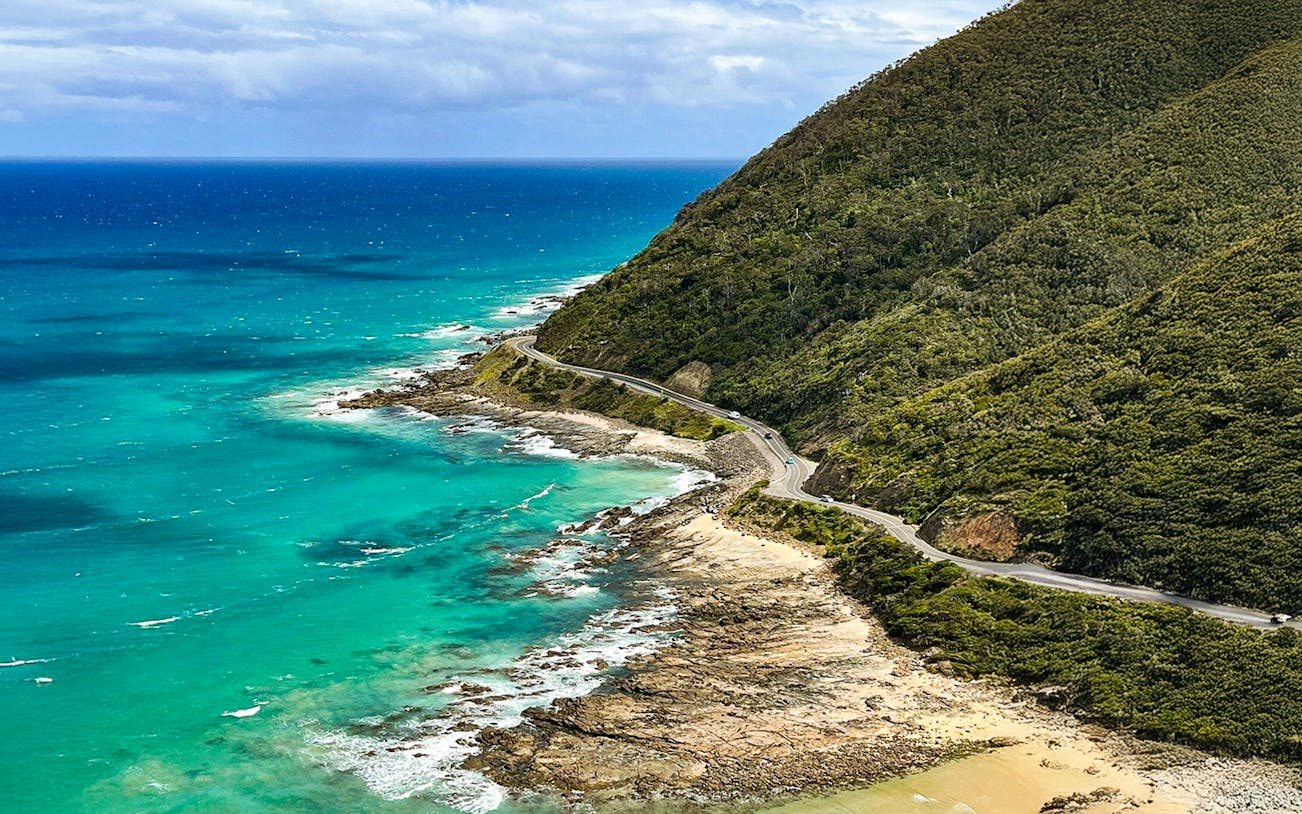 Aerial view of the Great Ocean Road winding along the coastline with turquoise waters and lush hills.