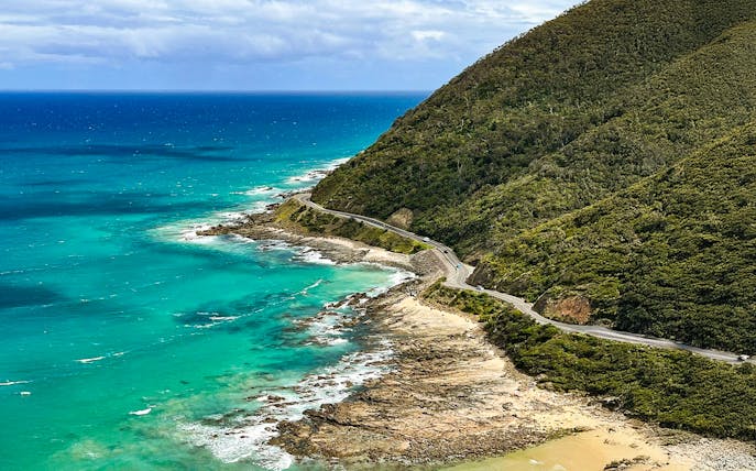 Aerial view of the Great Ocean Road winding along the coastline with turquoise waters and lush hills.