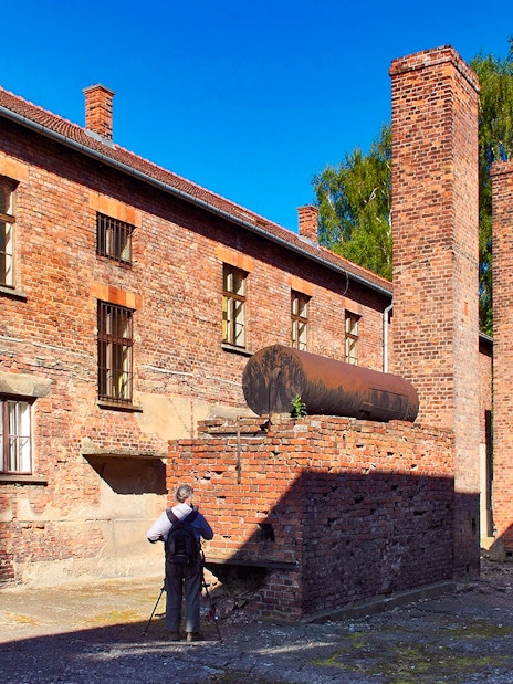 Barrack building and chimneys at Auschwitz Birkenau, Poland, with a visitor taking photos.