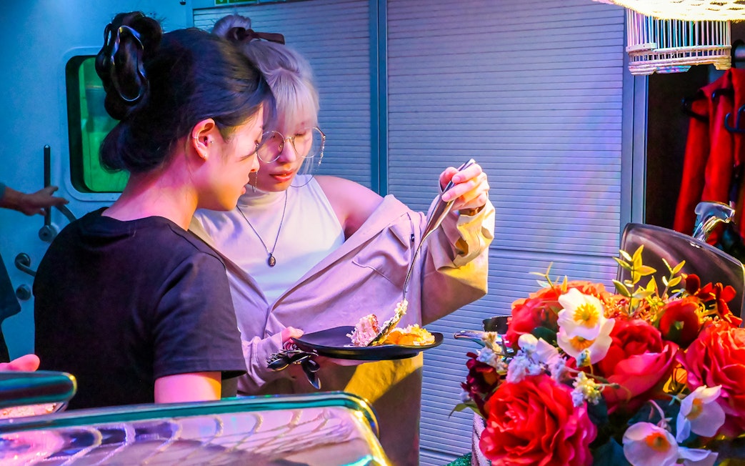 Two people selecting food at a vibrant market stall with flowers nearby.