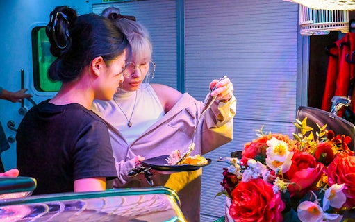 Two people selecting food at a vibrant market stall with flowers nearby.