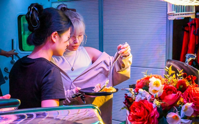 Two people selecting food at a vibrant market stall with flowers nearby.
