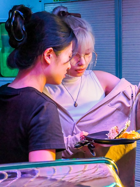 Two people selecting food at a vibrant market stall with flowers nearby.