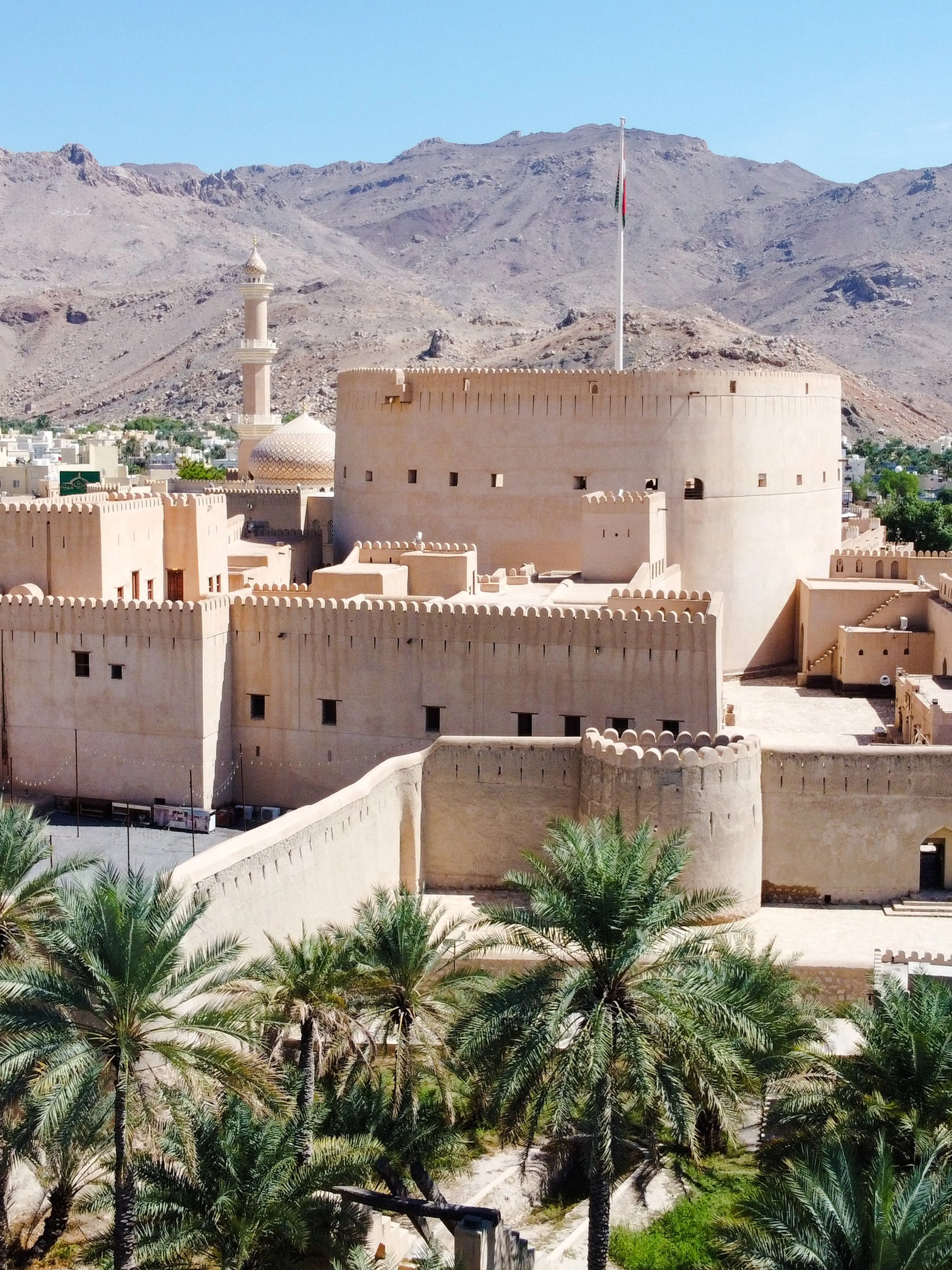 Aerial view of Nizwa Fort in Oman with surrounding mountains and palm trees.