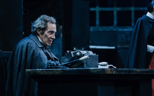 Man portraying Scrooge in a scene from "A Christmas Carol" at a desk in a dimly lit room.