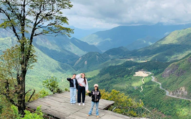 Tourists on a wooden platform overlooking Sapa's lush green mountains and winding roads.