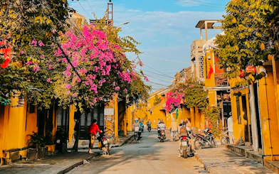 Hoi An street with yellow buildings and pink bougainvillea, motorbikes passing by.
