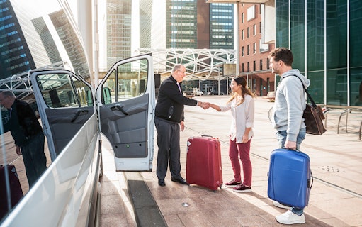 Airport shuttle driver greeting passengers with luggage outside a van.