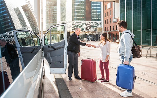 Airport shuttle driver greeting passengers with luggage outside a van.