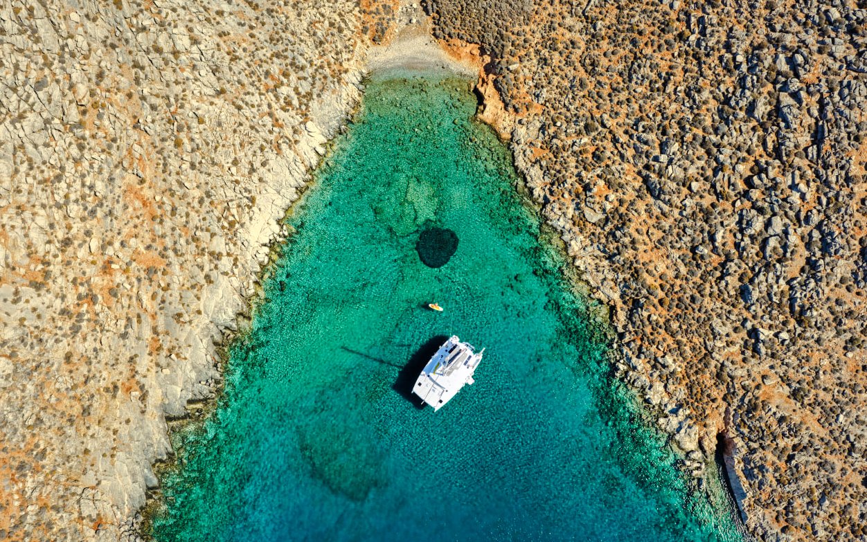 Aerial view of a catamaran in turquoise waters near rocky Dia Island coast.