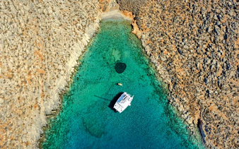Aerial view of a catamaran in turquoise waters near rocky Dia Island coast.