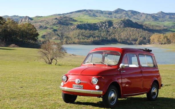 Red vintage car parked by a lake with hills in the background, Riviera of the Cyclops tour.