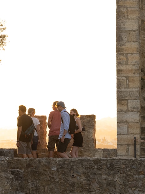 Visitors exploring St. George Castle at sunset, Lisbon, with audio tour on phone.