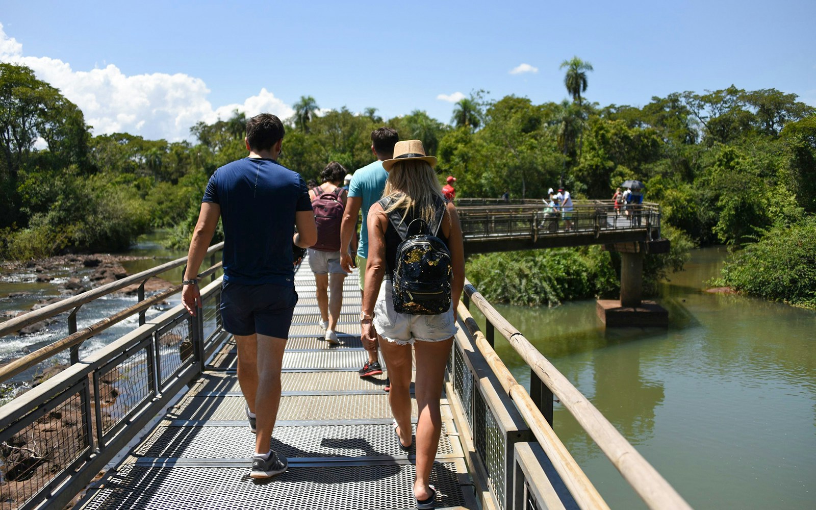 Visitors walking on a bridge towards Iguazu Falls, surrounded by lush greenery.