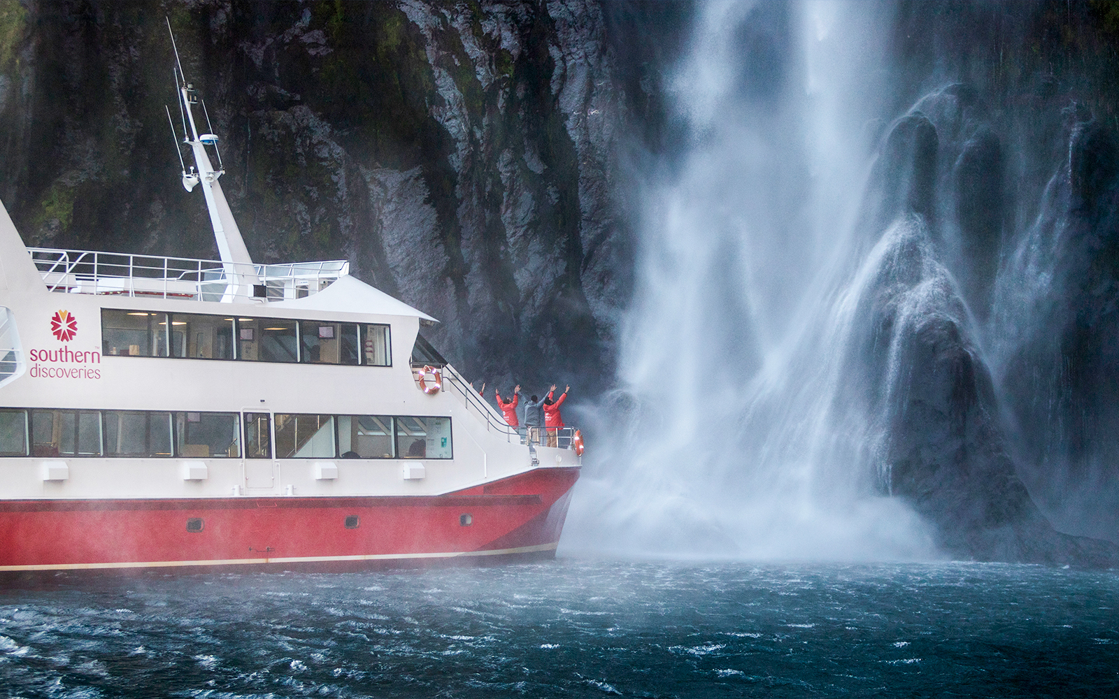 Cruise ship near waterfall in Milford Sound, New Zealand, with passengers on deck.