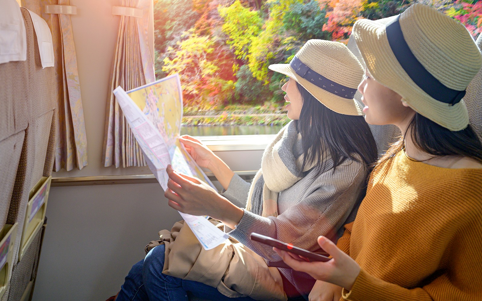 Two women in hats looking at a map inside a train with autumn scenery outside.
