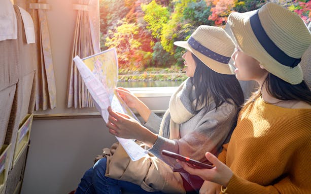 Two women in hats looking at a map inside a train with autumn scenery outside.