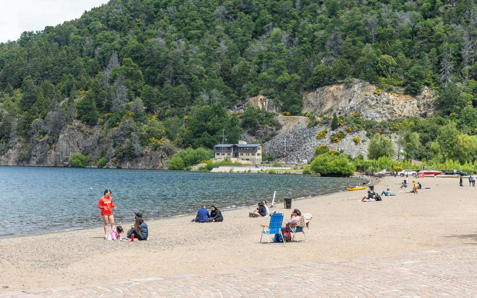 People relaxing on the sandy beach by Lake Lácar with forested hills in the background.
