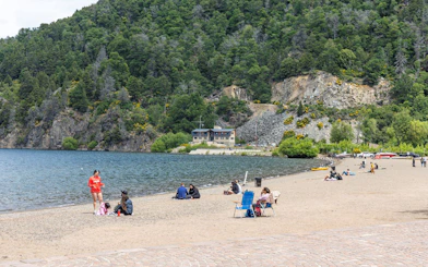 People relaxing on the sandy beach by Lake Lácar with forested hills in the background.