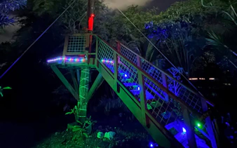 Nighttime zipline platform illuminated with colorful lights in a forest setting.