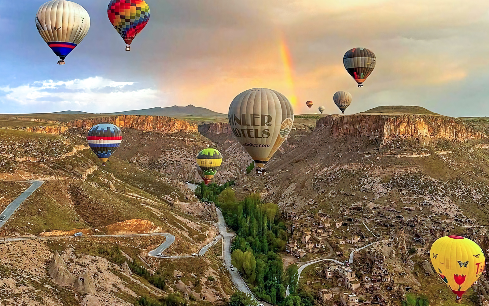 Hot air balloons floating over Soganli Valley, Cappadocia, with a rainbow in the background.
