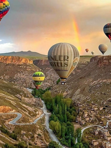 Hot air balloons floating over Soganli Valley, Cappadocia, with a rainbow in the background.