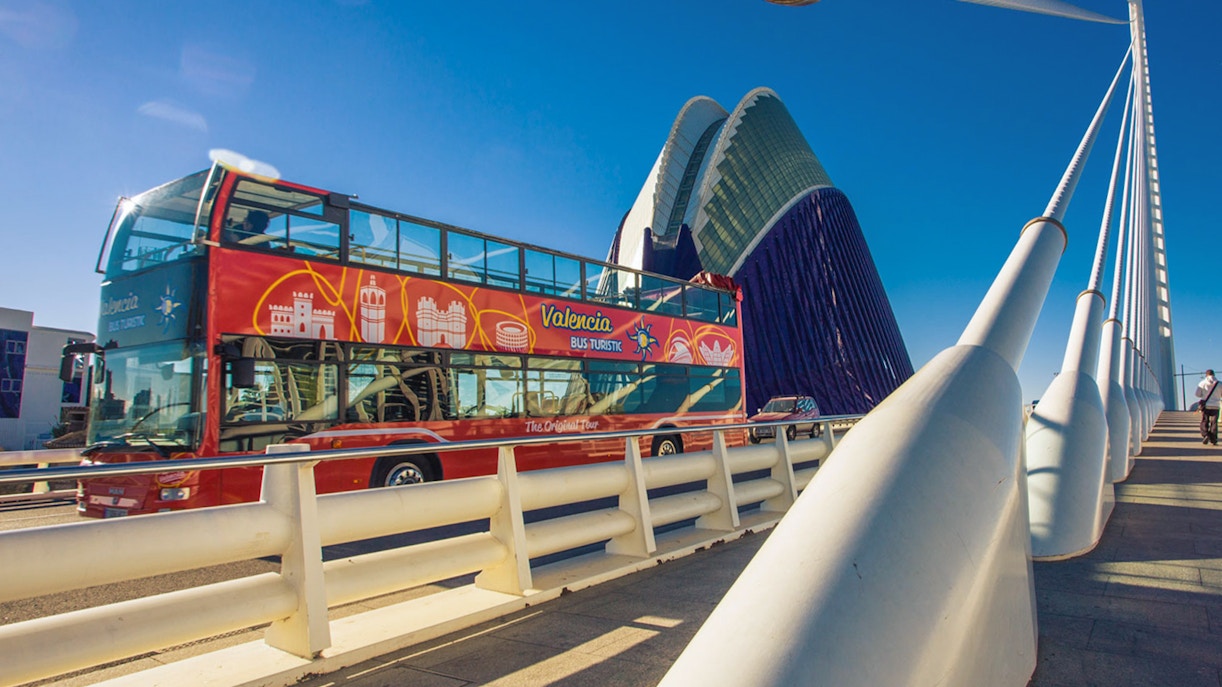 Valencia Hop On Hop Off bus crossing a bridge with cityscape in the background.