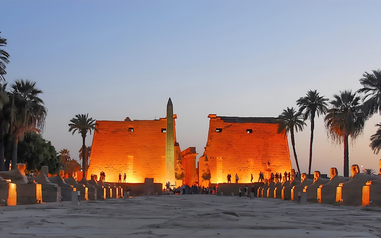 Luxor Temple entrance at dusk with illuminated obelisk and palm trees, Egypt.