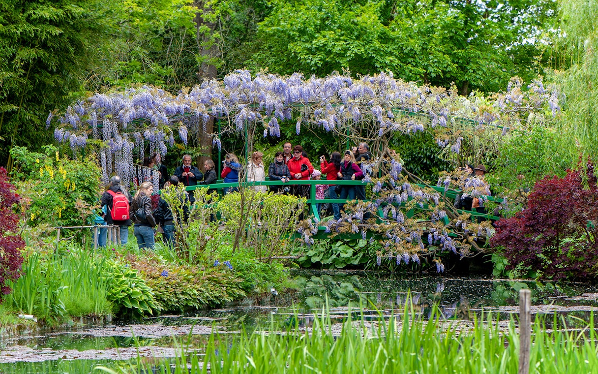Visitors on a bridge in Monet's Garden, Giverny, surrounded by wisteria and lush greenery.