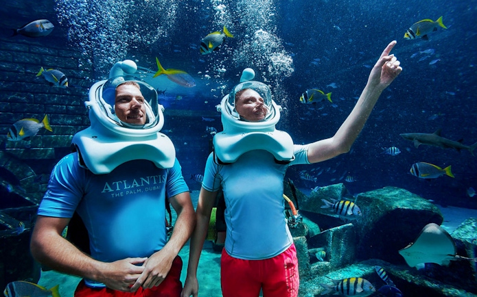 Tourists scuba diving with helmets at Atlantis, surrounded by fish and marine life.