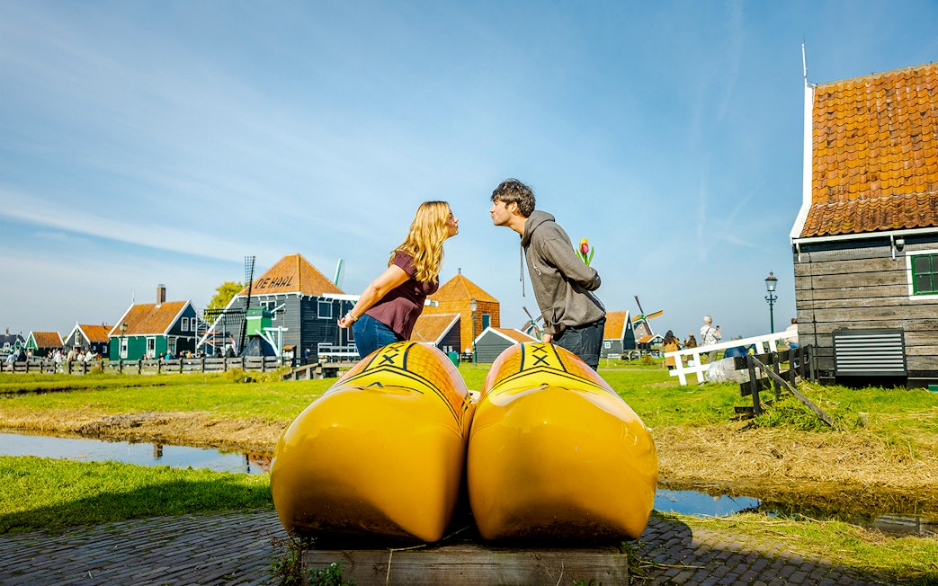 Guests at Zaanse Schans posing with large yellow clogs, traditional Dutch houses in background.