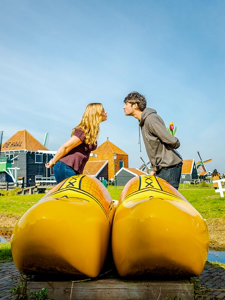 Guests at Zaanse Schans posing with large yellow clogs, traditional Dutch houses in background.