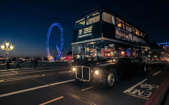 London double-decker bus on ghost tour passing the illuminated London Eye at night.