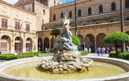 Fountain with statue in Monreale square, Sicily, with historic architecture in the background.