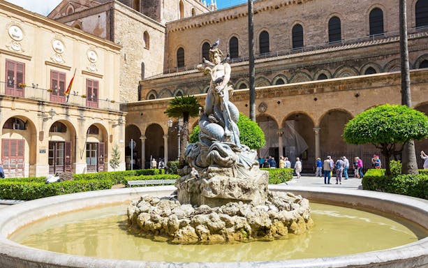 Fountain with statue in Monreale square, Sicily, with historic architecture in the background.