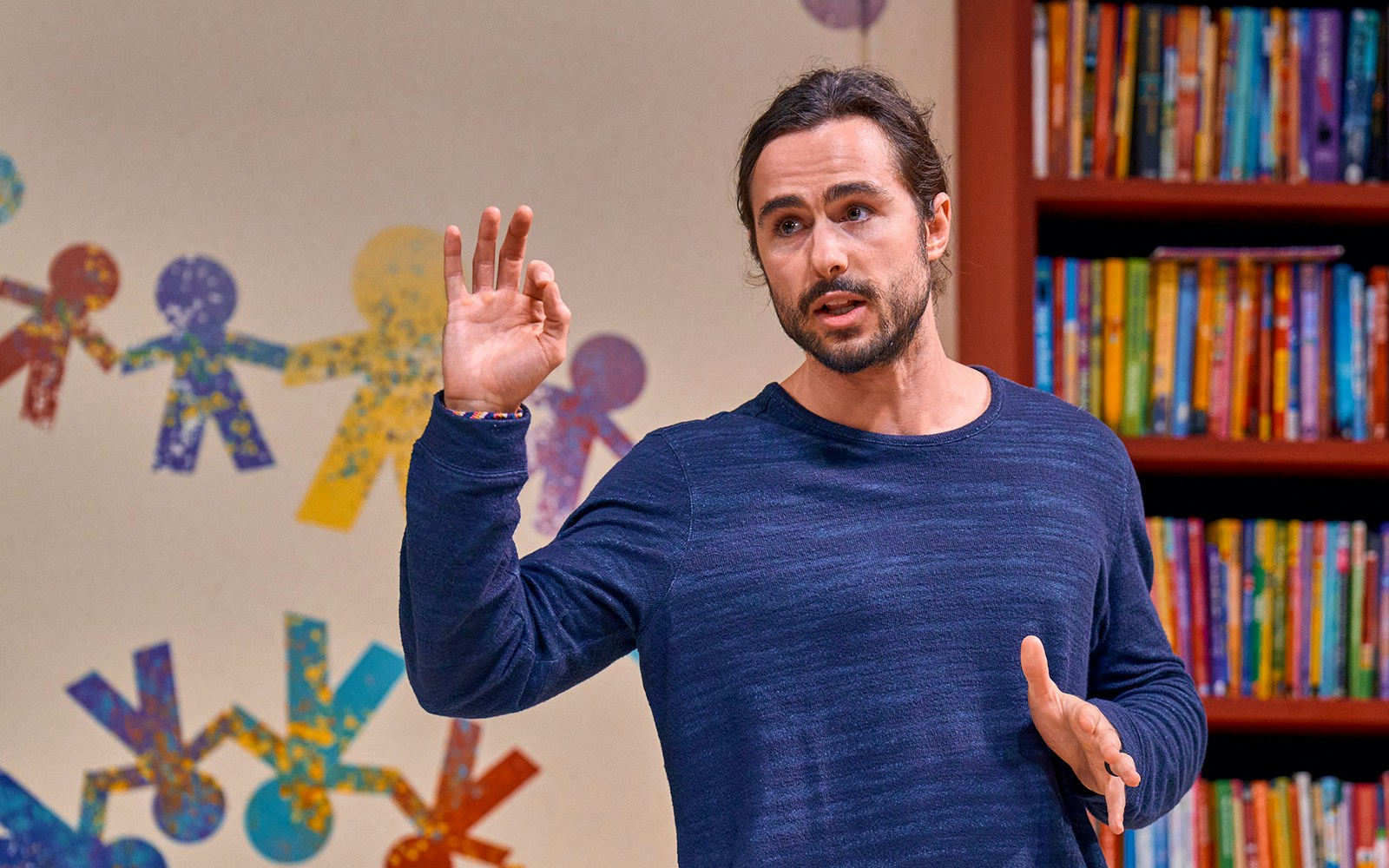 Man speaking in front of colorful paper chain figures and bookshelves during Eureka Day event.