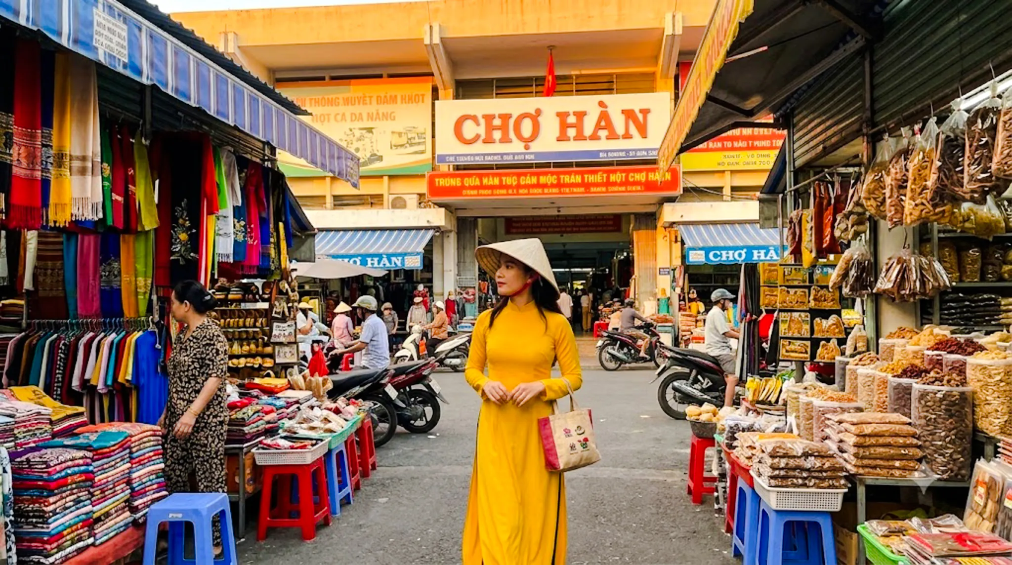 Woman in traditional dress at Chợ Hàn market, Da Nang, surrounded by colorful stalls and local goods.